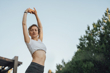 A girl in grey leggings and a white top raised her hands up looking down and doing a sporting exercise against the sky in the park's outdoor frame to waist