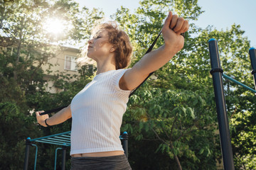 A woman in grey leggings and a white top looks in front of her and performs an exercise in her arms together to frame the waist with a sunny glare from behind