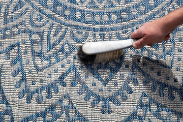 Man brushing an outdoor rug in a garden with a hand brush.  Grey and blue design