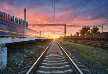 Fototapeta na wymiar Railway station with blurred high speed train and freight train at colorful sunset. Railroad in summer. Industrial landscape with moving train, railway platform, sky with pink clouds. Transportation