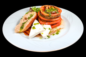 Gourmet, goat cheese salad with tomato, roasted zucchini, baguette and basil sauce on a white round plate, Isolated on a black background