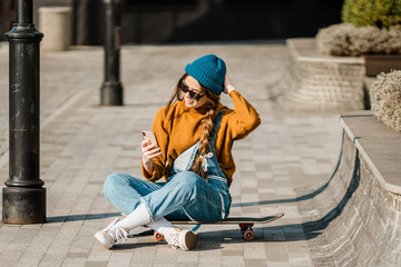 Cute urban girl outdoors with skateboard using smart phone. Happy young woman with skateboard, headphones and cell phone. Hipster female sit on skate board and using mobile phone