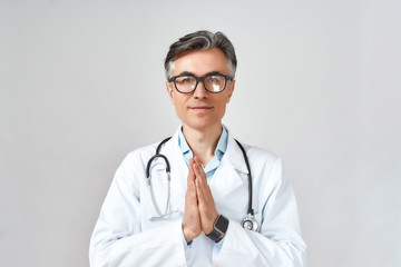Senior professional male doctor or physician in white coat with stethoscope around neck praying, looking at camera while standing against grey background