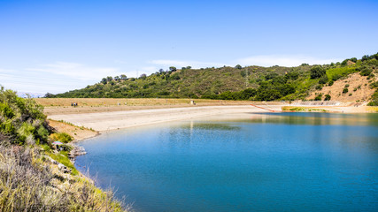 Fototapeta na wymiar Stevens Creek reservoir on a sunny day, with people walking and relaxing on the dam and shoreline; south San Francisco bay area, California