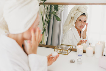 Obraz na płótnie Canvas Young beautiful woman wearing bathrobe and towel on her hair applying moistrizing cream on her face. Skin care morning rituals. Beauty routine. 