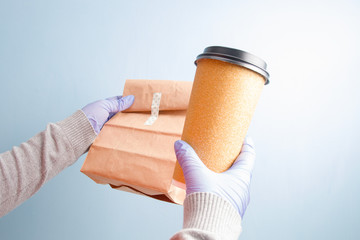 female hands in blue rubber disposable gloves hold a large paper cup of coffee and a paper bag, blue background, copy space, contactless delivery concept, focus on the bag