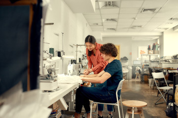 Obraz premium The Joy of Crafting Best. Young man, clothing designer sitting and sewing. His female colleague helping him. Dressmaker working on the sewing machine. Tailor making a garment in his workplace