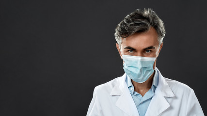 Protective equipment. Confident mature male doctor wearing medical uniform and protective mask looking at camera while standing against black background