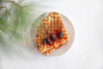 waffles with jam on a white saucer on the table and a glass vase with a pine branch