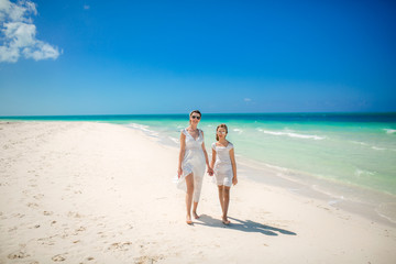 Mother and daughter in white dresses walking on the beach. Family by the ocean in sunglasses.