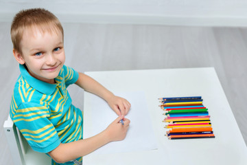 A beautiful boy draws a cute drawing with colored pencils for fathers ' Day. A cute child is sitting at a table and holding a pencil in his hand. Elementary school and homeschooling concept.