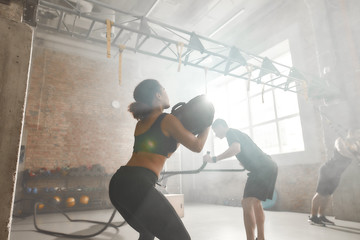 Fototapeta na wymiar Live in your Passion. Sportive woman lifting a heavy sandbag while having workout at industrial gym. Group training, teamwork concept