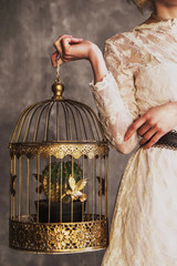 woman in a white lace dress holding a golden cage with gold birds and a cactus. no face psychology therapy concept. women empowerment, golden cage syndrome. entrapment Stockholm syndrome. girl power
