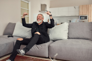 Adding Fun to Life. Full-length shot of bearded middle-aged man excited about winning while playing video games after smoking marijuana from a bong or glass water pipe sitting on the couch at home