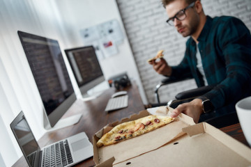 Time for a break. Young male web developer in a wheelchair eating pizza while sitting at his workplace in the modern office. Disabled male programmer having a lunch