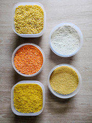 Five plastic containers with dry food (rice, red lentils, vermicelli pasta, flour and couscous) displayed on wooden table. Top view, from above, flat lay.