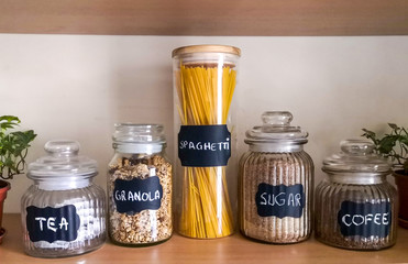 Five glass jars containing tea, granola, spaghetti, sugar and coffee sitting on a wooden shelf on the wall with two English ivy plants on both sides.
