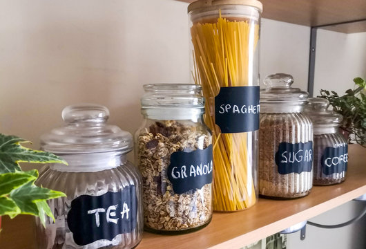 Five glass jars containing tea, granola, spaghetti, sugar and coffee sitting on a wooden shelf on the wall with two English ivy plants on both sides.

