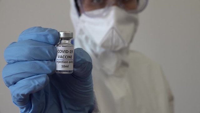 Doctor in protective PPE suit and medical gloves shows small coronavirus vaccination bottle. Answer to rapidly spreading global Covid-19 pandemic.
