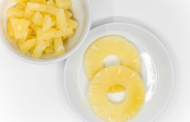 slices of canned pineapple on a white plate and chopped pineapple in a white porcelain bowl on a white background