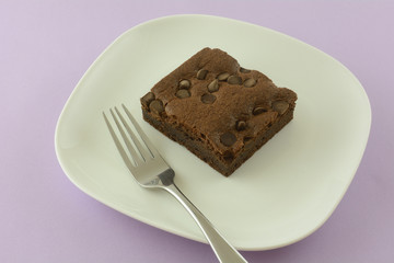 Chocolate chip brownie square on white dessert plate with fork on lavender background