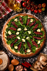 Tasty arugula and dried tomato pizza and cooking ingredients tomatoes basil on wood background. Top view of arugula pizza