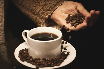 Mano blanca dejando caer un puñado de granos de café tostado sobre un plato blanco con taza de café sobre fondo negro.