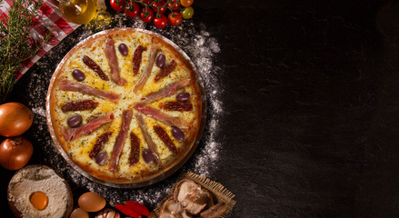 Tasty prosciutto pizza and cooking ingredients tomatoes basil on black concrete background. Top view prosciutto pizza