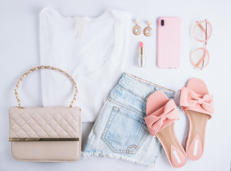 Female clothes and accessories with white t-shirt, jeans, sunglasses, lipstick, pink shoes, earrings and smartphone on white background. Flat lay.