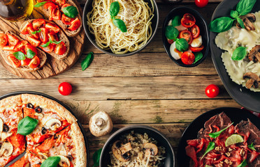 Table of italian meals on plates Pizza, pasta, ravioli, carpaccio, mushroom risotto, caprese salad and tomato bruschetta on rustic wooden background.