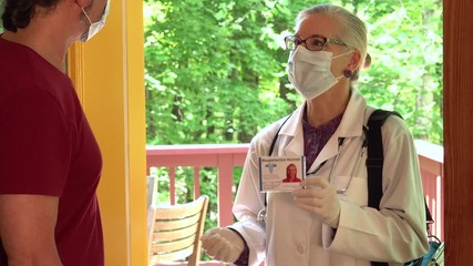Closeup of mature woman nurse or doctor wearing face mask at home door showing badge, greeting, and entering.