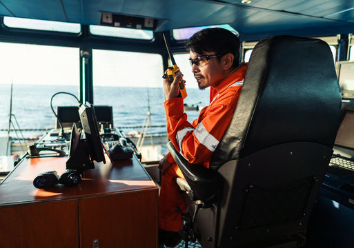 Filipino deck Officer on bridge of vessel or ship wearing coverall during navigaton watch at sea . He is speaking on portable GMDSS VHF radio, short range communication