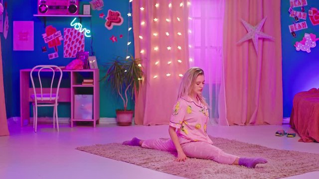 Beautiful blonde young woman practicing sport in her room on the carpet she stretching her legs while wearing a cute pajama in the morning