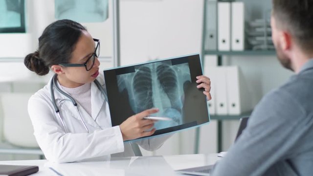 Female doctor in lab coat and glasses pointing at lung abnormality on x-ray scan and explaining diagnosis to male patient during medical consultation in clinic. Covid-19 outbreak concept