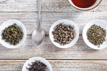 A top down view of several varieties of dried loose leaf teas on small saucer plates, on a wooden table surface.