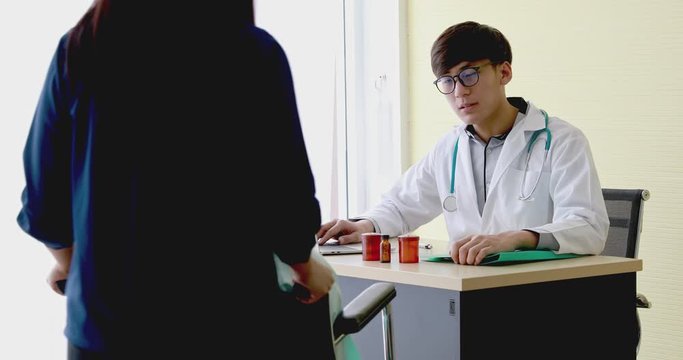 Young asian doctor with glasses talking and consulting with female patient on wheelchair about illness and recovery with smiley and confident look while making note on his file at hospital