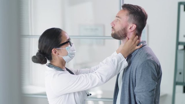 Female Doctor In Protective Face Mask Touching Lymph Nodes On Neck And Examining Throat Of Male Patient During Medical Checkup In Clinic