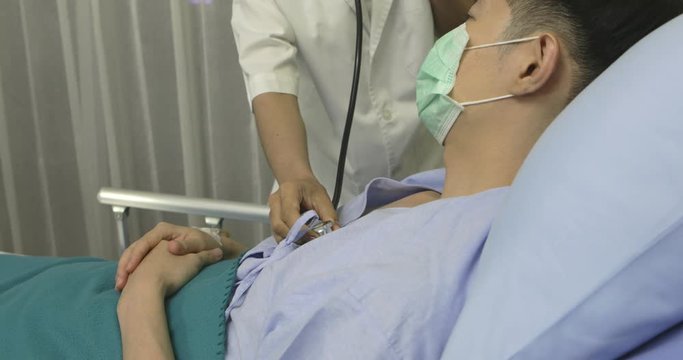 Close-up of asian male dotor use stethoscope to examing on young asian patient wearing surgical mask on his chest in hospital bed.