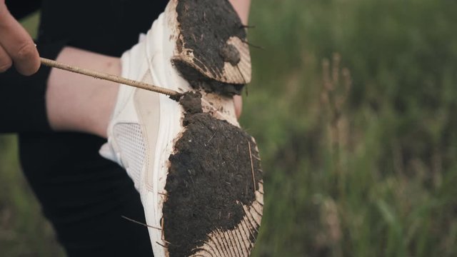 Girl in a white shirt and white sneakers cleans the dirt from the sole while standing on one leg, close up