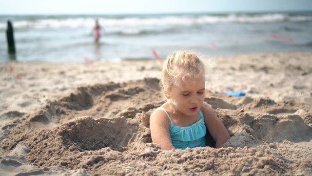 Blond adorable girl buried in sand try to break free. Blurred people and sea