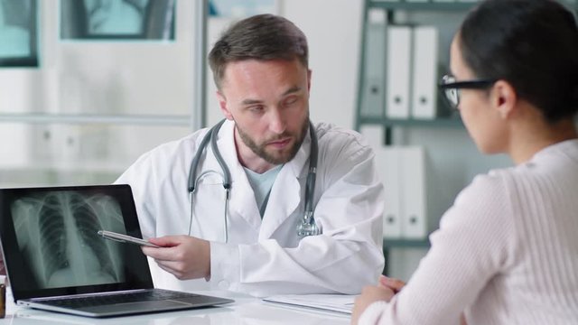 Professional male doctor in lab coat showing lung x-ray on laptop screen and explaining diagnosis to female patient during consultation in medical office