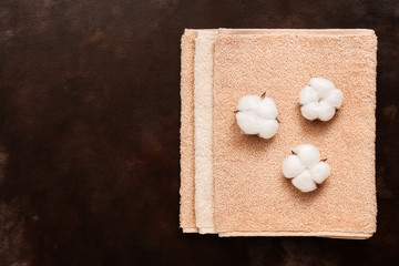 Set of beige terry towels and cotton flowers on a dark brown background. Top view, flat lay, copy space.