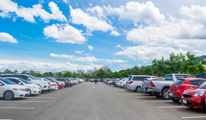 Car parking in large asphalt parking lot with trees, white cloud and blue sky background.