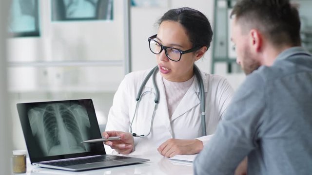 Young mixed-raced female doctor in lab coat and glasses pointing at lung x-ray on laptop screen and explaining diagnosis to male patient during medical consultation in clinic