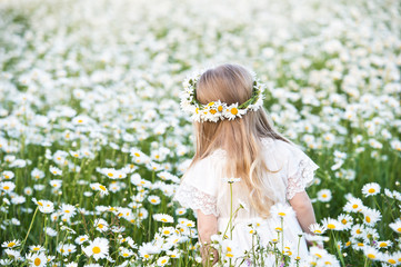 Beautiful girl with blond hairand chamomilie wreath in chamomile field. Portrait of cute little girl child with bouquet of chamomiles flowers in sunny summer day.