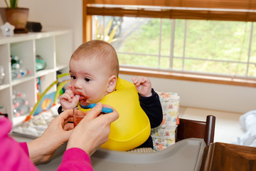 Adorable little baby boy in feeding chair being spoon fed by his mother