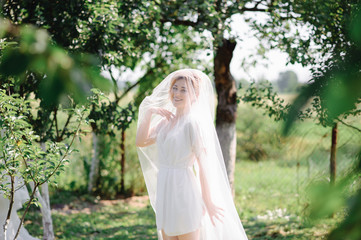 Fototapeta na wymiar Beautiful blonde bride posing under a veil in a bathrobe in a green garden.