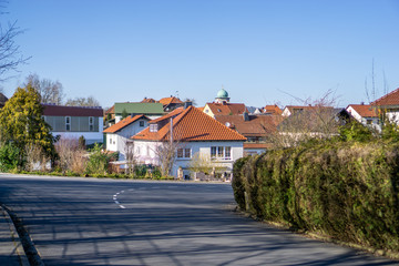 Leere Straße in einer Wohnsiedlung macht einen Bogen entlang einer Hecke. Im Hintergrund sieht man die Spitze eines Kirchturms und blauen Himmel. Landleben bei Tageslicht.