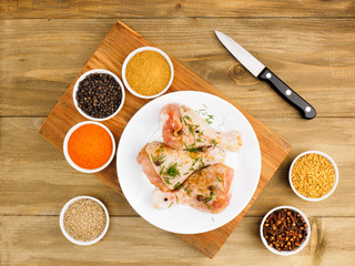 Raw chicken drumsticks on white ceramic plate, knife, spice set red chili pepper, masala, black pepper, cumin, fenugreek, fennel, cloves