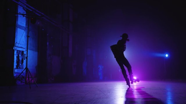 Rear shot of professional roller skater wearing cap and shirt riding around stage under blue and violet lighting and doing various complex tricks and turns. Experienced roller skater performance